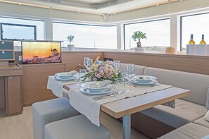 A dining area with a table set for four, a television displaying a sunset scene, and large windows offering a view of the sky. - Sun-Kissed Sailing Adventure in Islas Sarónicas (Alimos)