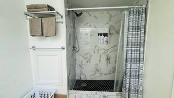Eine Dusche mit Glastür, ein Handtuchhalter mit Handtüchern, ein Duschkopf und eine kleine Ablage mit Pflegeprodukten.