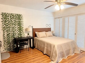 Quarto com cama, escrivaninha, cadeira, ventilador de teto e parede com folhagem verde.