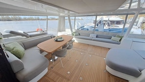 A yacht interior with a wooden deck, a sofa, a table, and a view of the marina. - Sun-Kissed Sailing Adventure in Bonifacio, Córcega (Bonifacio)