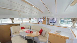 A boat interior with a dining area, cushioned seating, a small table with a bottle, and windows with curtains. - Sun-Kissed Sailing Adventure in Islas Sarónicas (Alimos)