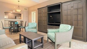 Een woonkamer met een bank, fauteuils, een salontafel, een televisie en een keuken op de achtergrond.