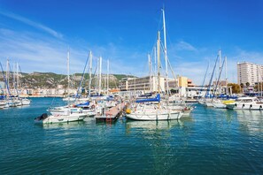 A marina with multiple boats docked, a building with a flat roof, and a hilly landscape in the background.