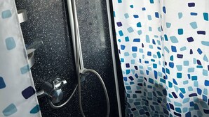 Dutxa amb vàlvula de pluja manual i plat de dutxa negre amb puntets, i cortina de dutxa blanca amb motius de mosaic blau.