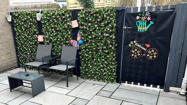 Zona de pati amb espai per seure, paret decorativa plena de flors i una porta amb un cartell de benvinguda.