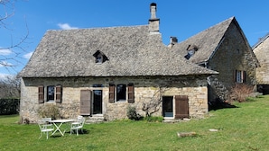 Een stenen huis met schuin dak, schoorsteen en een kleine tuin met een tafel en stoelen.