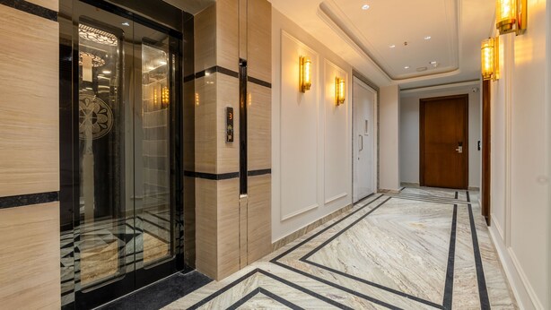 A hallway with a marble floor, glass elevator doors, wall sconces, and a wooden door at the end. - Anu Plaza-5Mins from Connaught Place (New Delhi)