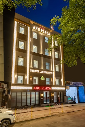 Un hotel de varios pisos llamado 'ANU PLAZA' con señalización iluminada, una entrada destacada y un coche estacionado en la parte delantera.