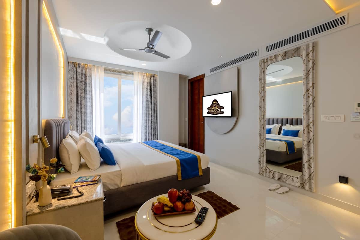 A modern hotel room with a bed, a desk with a vase and flowers, a fruit bowl, a remote control, and a large mirror.