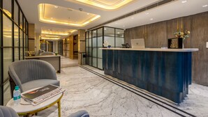 A hotel reception area with a marble floor, a reception desk, a grey armchair, and a small table with a magazine and a water bottle. - Anu Plaza-5Mins from Connaught Place (New Delhi)
