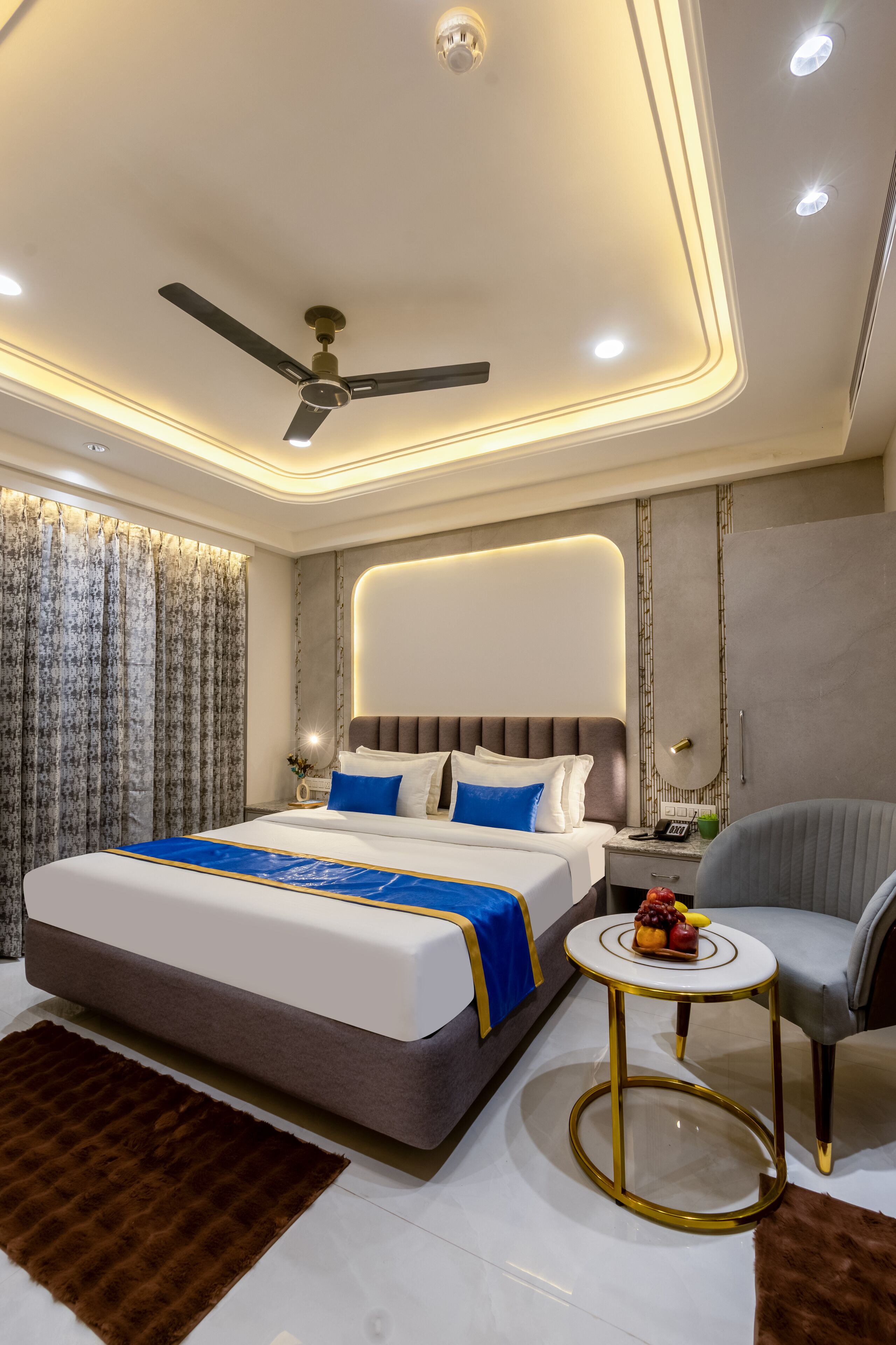 Una habitación de hotel moderna con una cama grande, un ventilador de techo, una mesita redonda con marco dorado y un sillón gris.