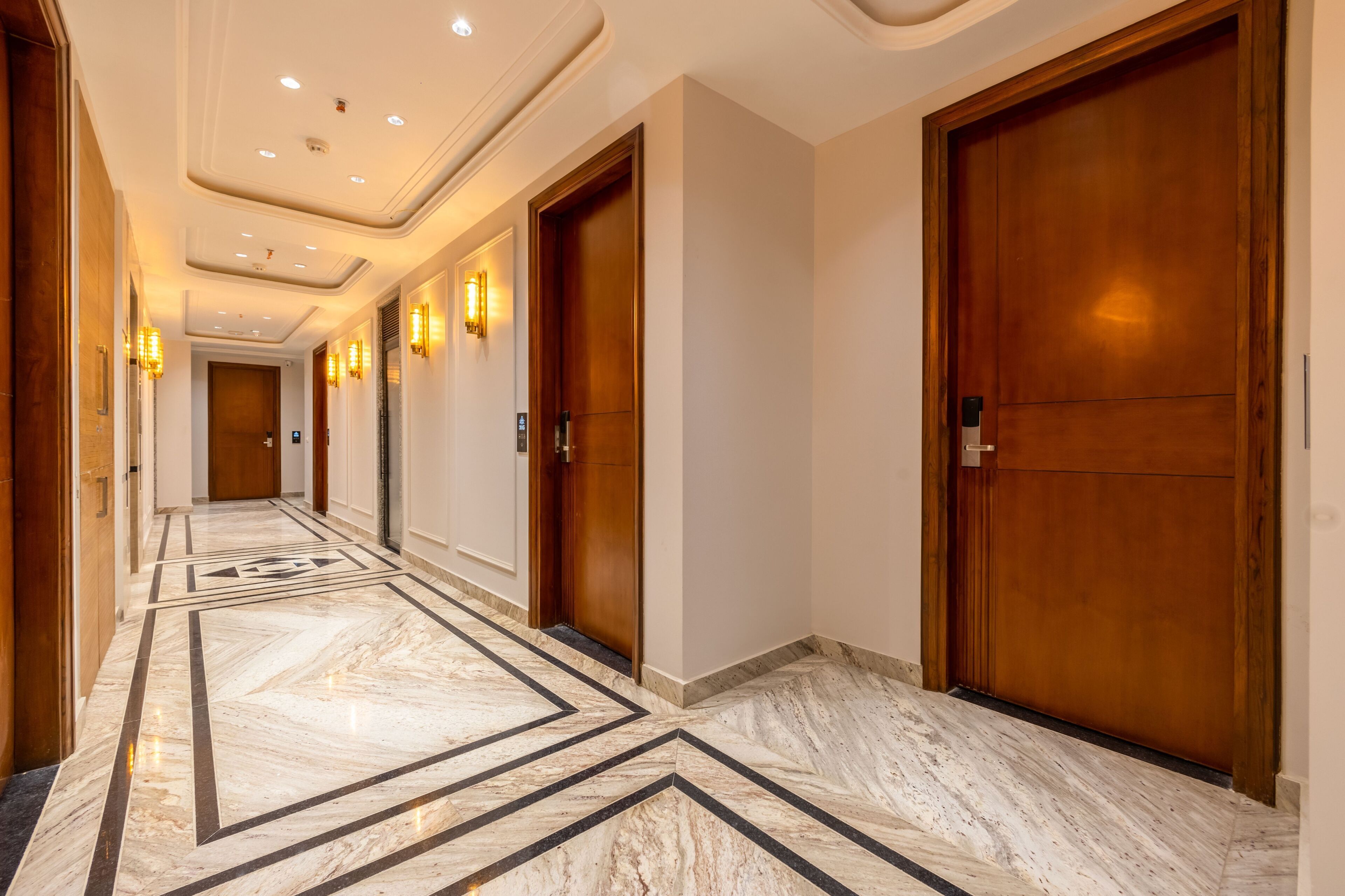 A hotel corridor with marble flooring, wooden doors, and wall-mounted lighting.