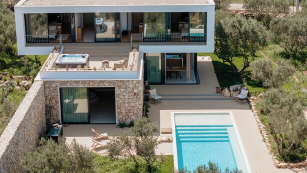 Una casa moderna de dos pisos amb una piscina, mobiliari d'exterior i una paret de pedra.