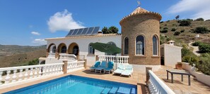 A poolside area with a clear view of a hillside, a building with a round tower, and solar panels on the roof. - Amazing Apartment in Algarrobo Campo with Views & Terrace (Algarrobo)