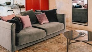 Una stanza con soffitto in legno, un divano grigio con cuscini, un mobiletto rosso, uno specchio e un pavimento piastrellato.