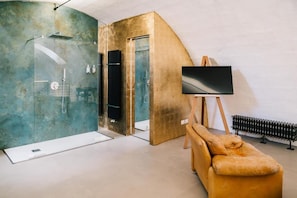 A modern bathroom with a glass shower enclosure, a golden accent wall, and a large TV on a wooden easel. - Il Granile Contemporary Loft  (Matera)