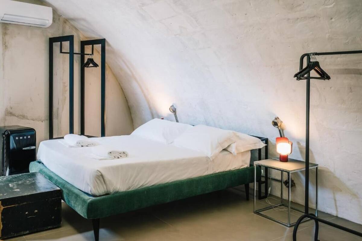 Una habitación con cama, una mesita de noche con lámpara, una maleta y un aire acondicionado instalado en la pared.
