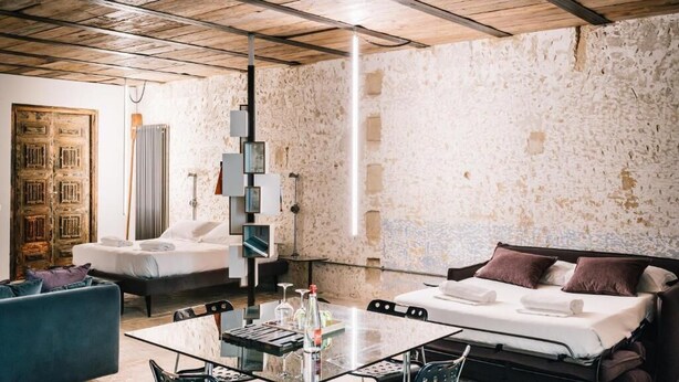 Una stanza con un letto, una poltrona, un tavolino in vetro e bottiglie, e un muro in pietra.