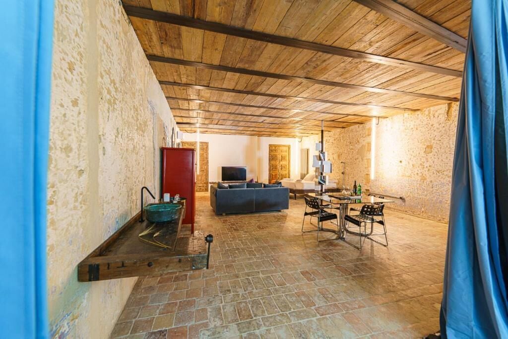 Un intérieur avec un mur en pierre, des poutres en bois au plafond, un canapé, un téléviseur et un coin repas avec des chaises et une table.