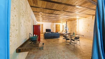 Un intérieur avec un mur en pierre, des poutres en bois au plafond, un canapé, un téléviseur et un coin repas avec des chaises et une table.