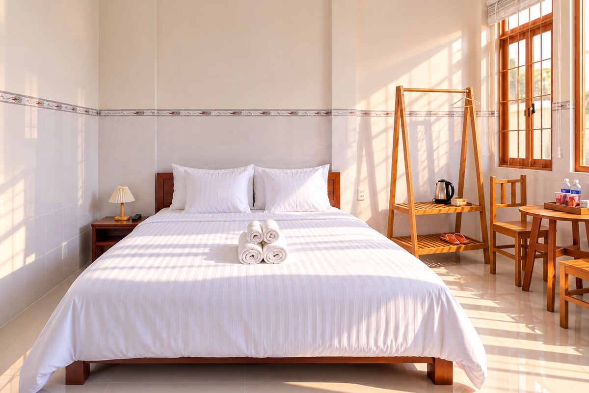 A hotel room with a bed, a wooden chair, a small table, and a window with blinds.