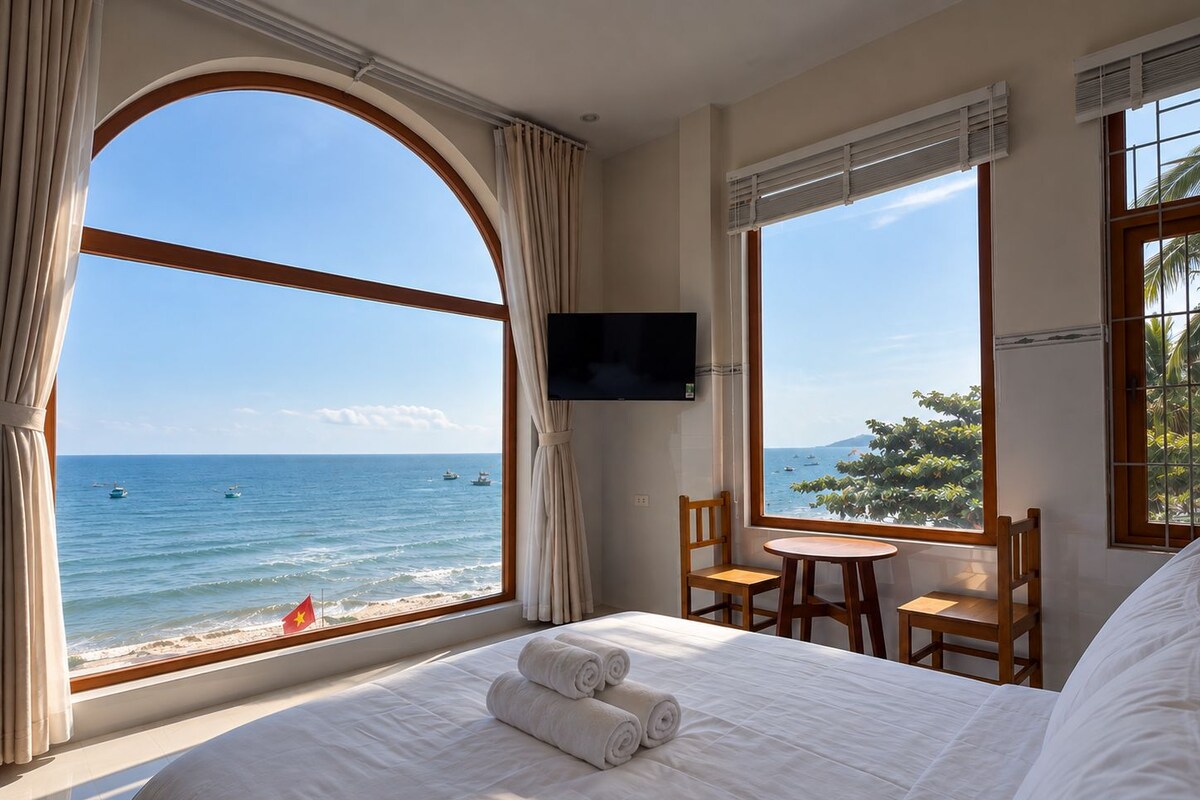 A hotel room with a large window offering a view of the sea, a bed with white linens, a television mounted on the wall, and a small table with chairs.