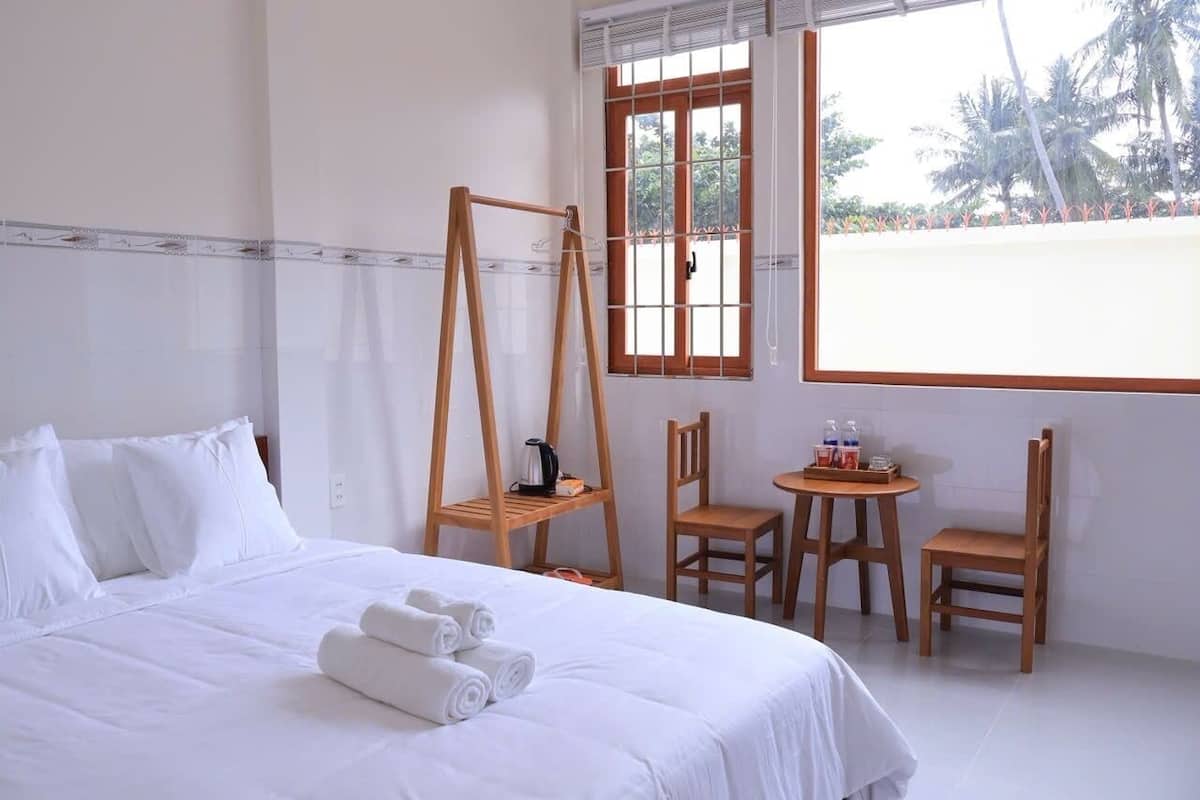 Habitación de hotel con cama, dos sillas de madera, una mesita con tetera y tazas, y una ventana con vistas a palmeras.