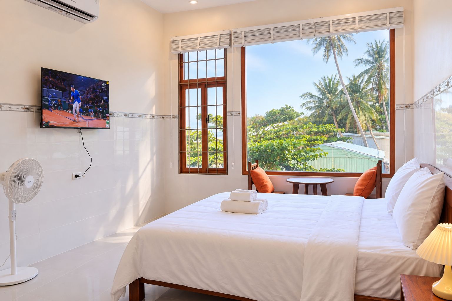 A hotel room with a bed, a TV, a fan, a window with a view of palm trees, and a small table with a chair.