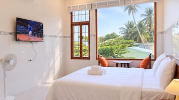 A hotel room with a bed, a TV, a fan, a window with a view of palm trees, and a small table with a chair.