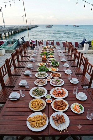 A long wooden dining table set with various dishes and drinks, overlooking a body of water with boats.