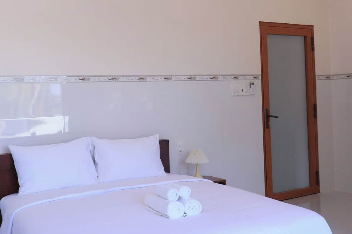 Una cama bien hecha con sábanas blancas y cabecera de madera, una lámpara de noche y una puerta con espejo.