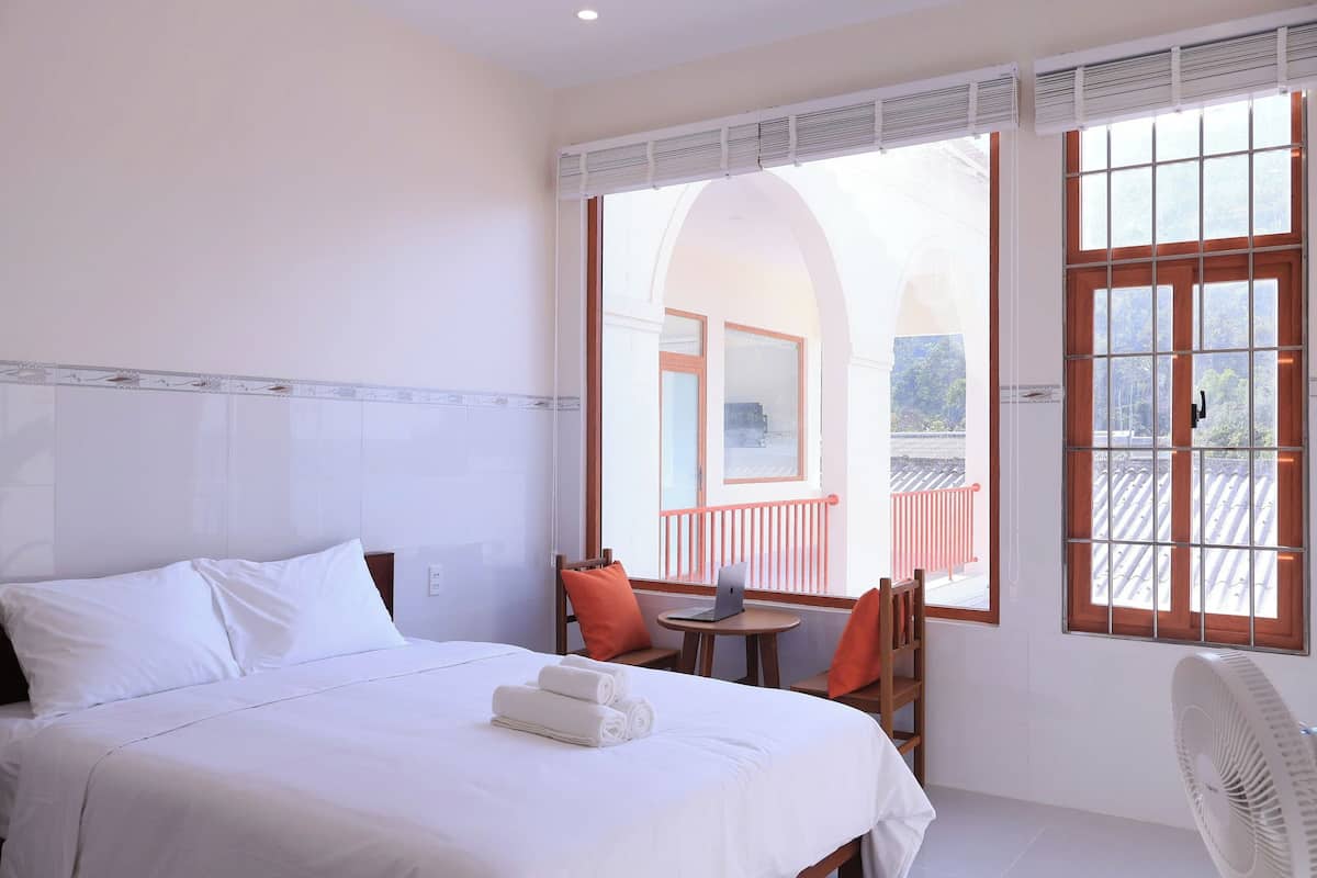 Habitación de hotel con cama, escritorio, silla, ventilador y ventanas amplias con vistas a un balcón.