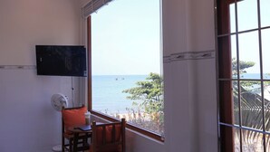 A room with a large window, a flat-screen TV mounted on the wall, a fan, and a chair with an orange cushion. - Prospera - Hòn Sơn Beachfront Hotel  (Kien Hai)