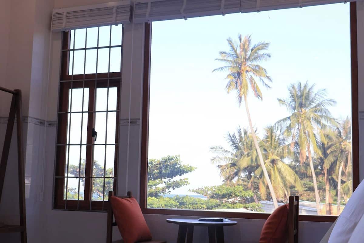 Habitación de hotel con cama, una mesita, dos sillas y ventana con vista a palmeras.
