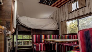 A compact RV interior with a bunk bed, a small dining area, and a window with a view of greenery. - 1 Bed Vehicle Luxury Camper -hot Tub, Pets - Willy (Skegness)