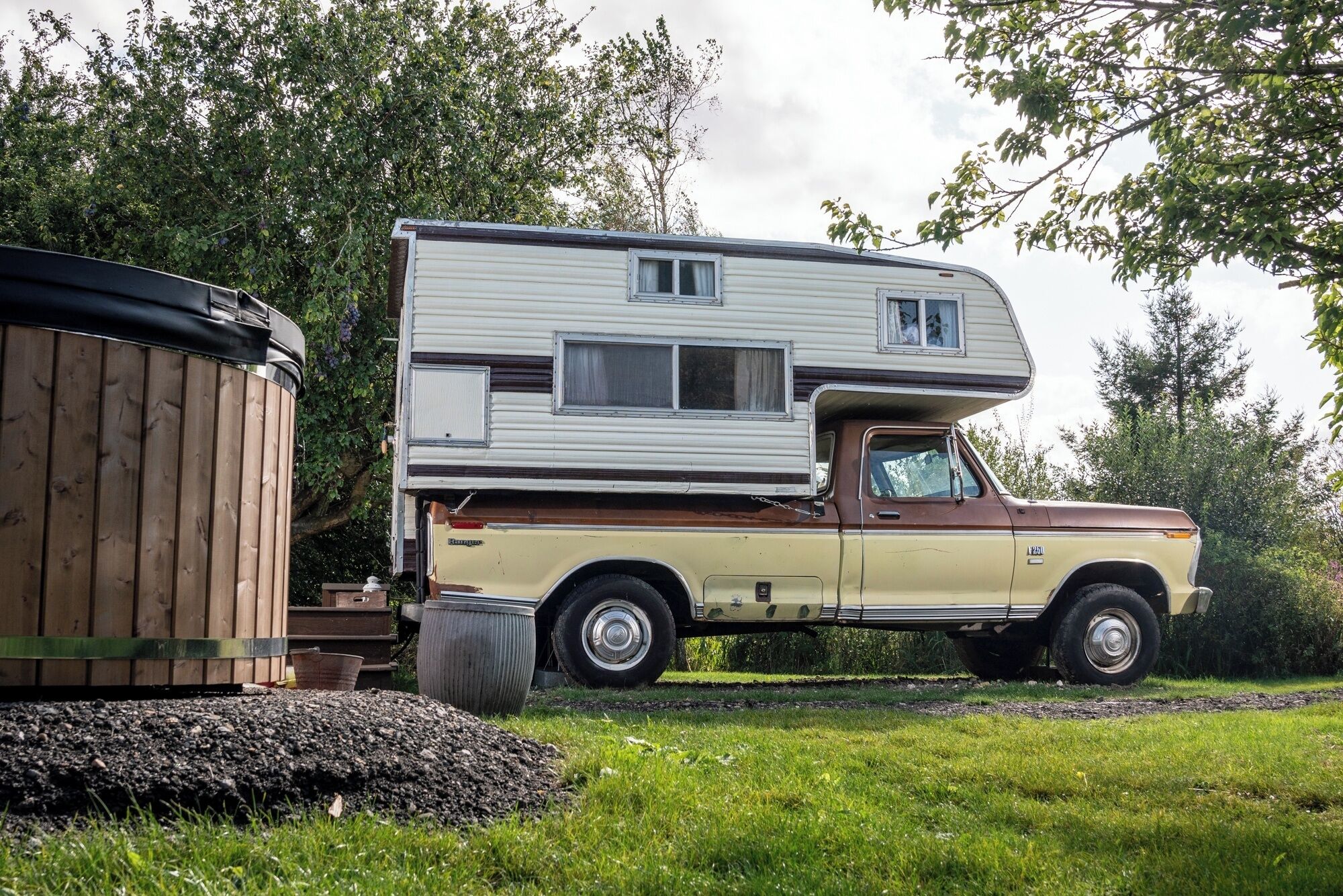 A vintage camper van parked next to a wooden structure, likely a storage shed, with a grassy area and trees in the background.