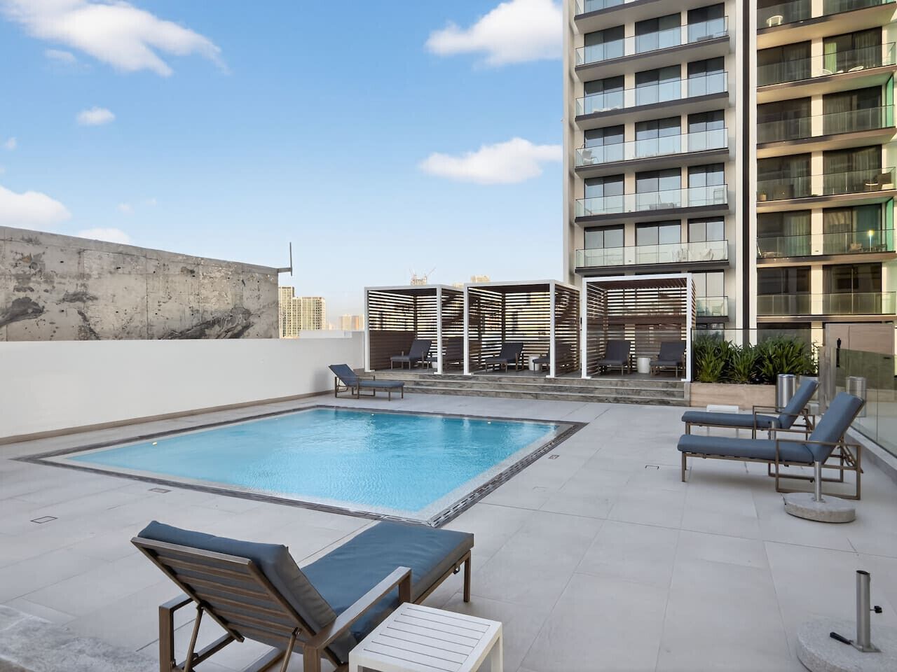 A rooftop pool with lounge chairs, a pergola, and a high-rise building in the background.