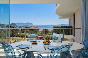 Een balkon met een eettafel gedekt voor twee personen, een fruitschaal en uitzicht op zee en bergen.