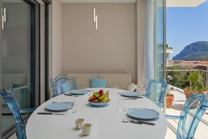 Een eetruimte met een ronde tafel, stoelen, een fruitschaal en uitzicht op zee en bergen.