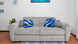 Ein graues Sofa mit zwei blau-schwarz gemusterten Kissen, einem Rahmenbild darüber und einer Pflanze zur Linken.