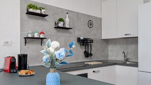 Eine moderne Küche mit grauer Arbeitsplatte, weißen Schränken, einer roten Kaffeemaschine, einer blauen Vase mit Blumen sowie einem Regal mit Pflanzen und Küchenutensilien.