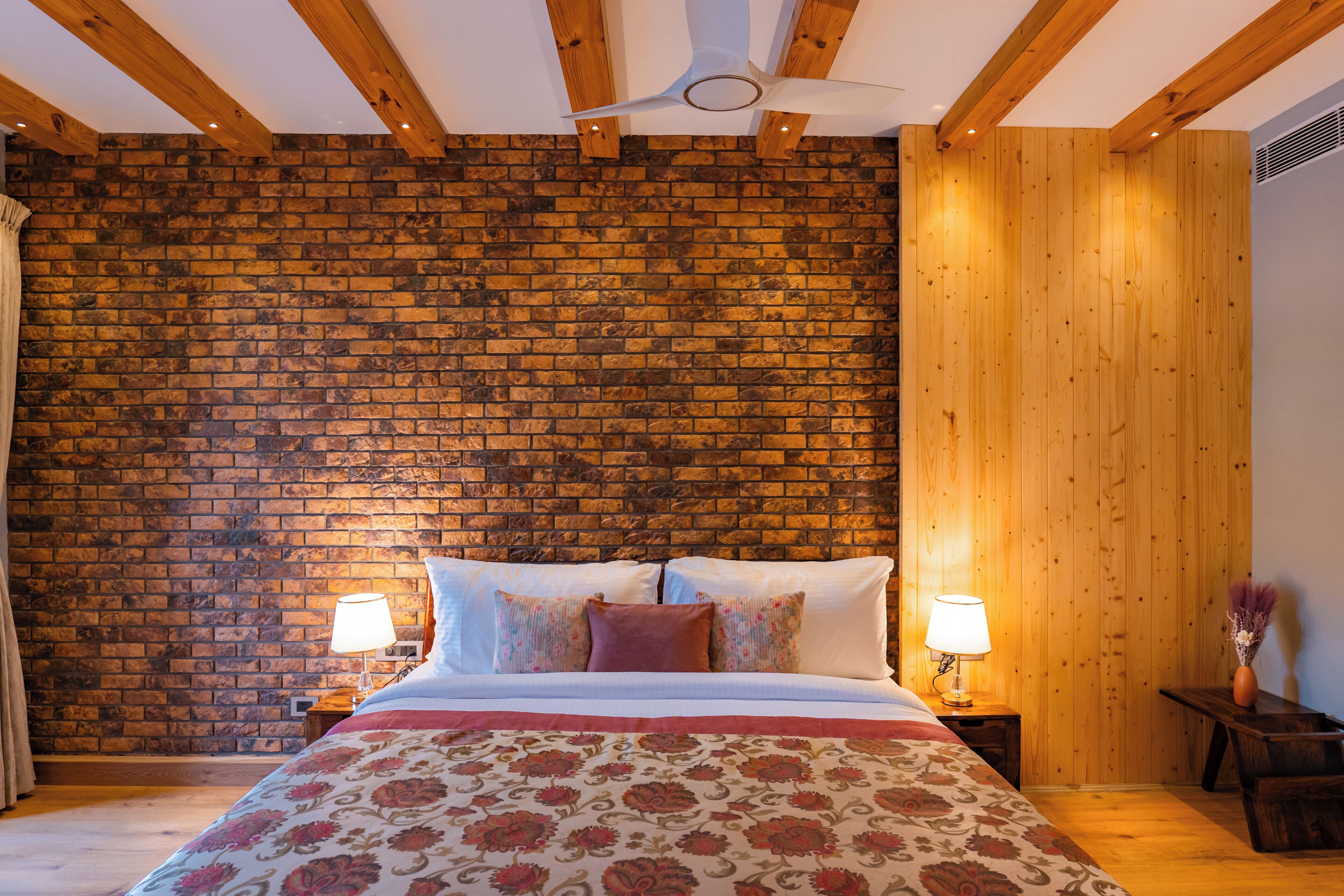 Een slaapkamer met een bakstenen muur, houten balken in het plafond, een bed met bloemenbeddengoed, twee nachtkastjes met lampen en een kleine tafel met een vaas.