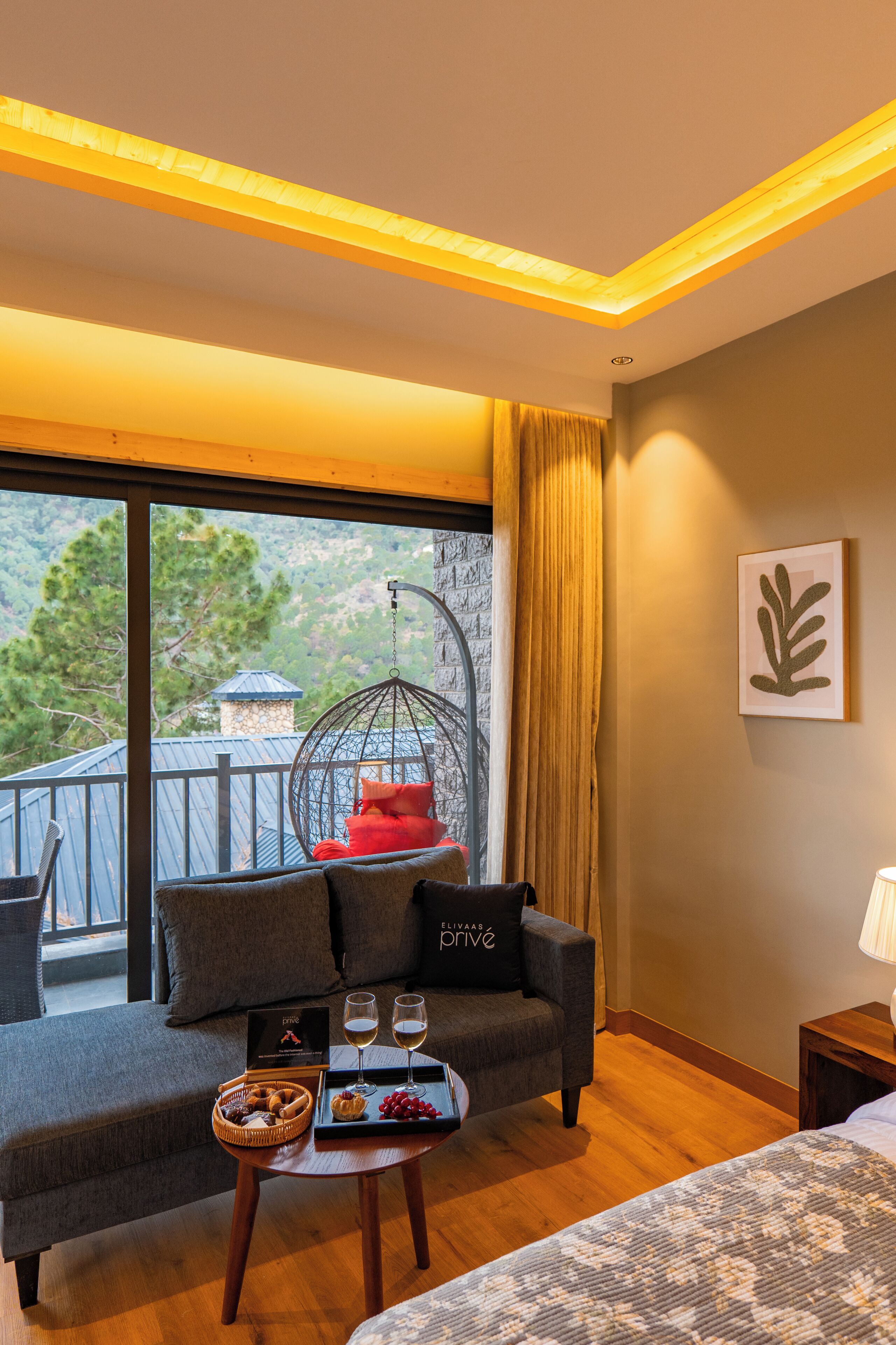 Quarto de hotel com varanda, sofá, cadeira suspensa, cama e mesa de jantar com bandeja de petiscos e copos.
