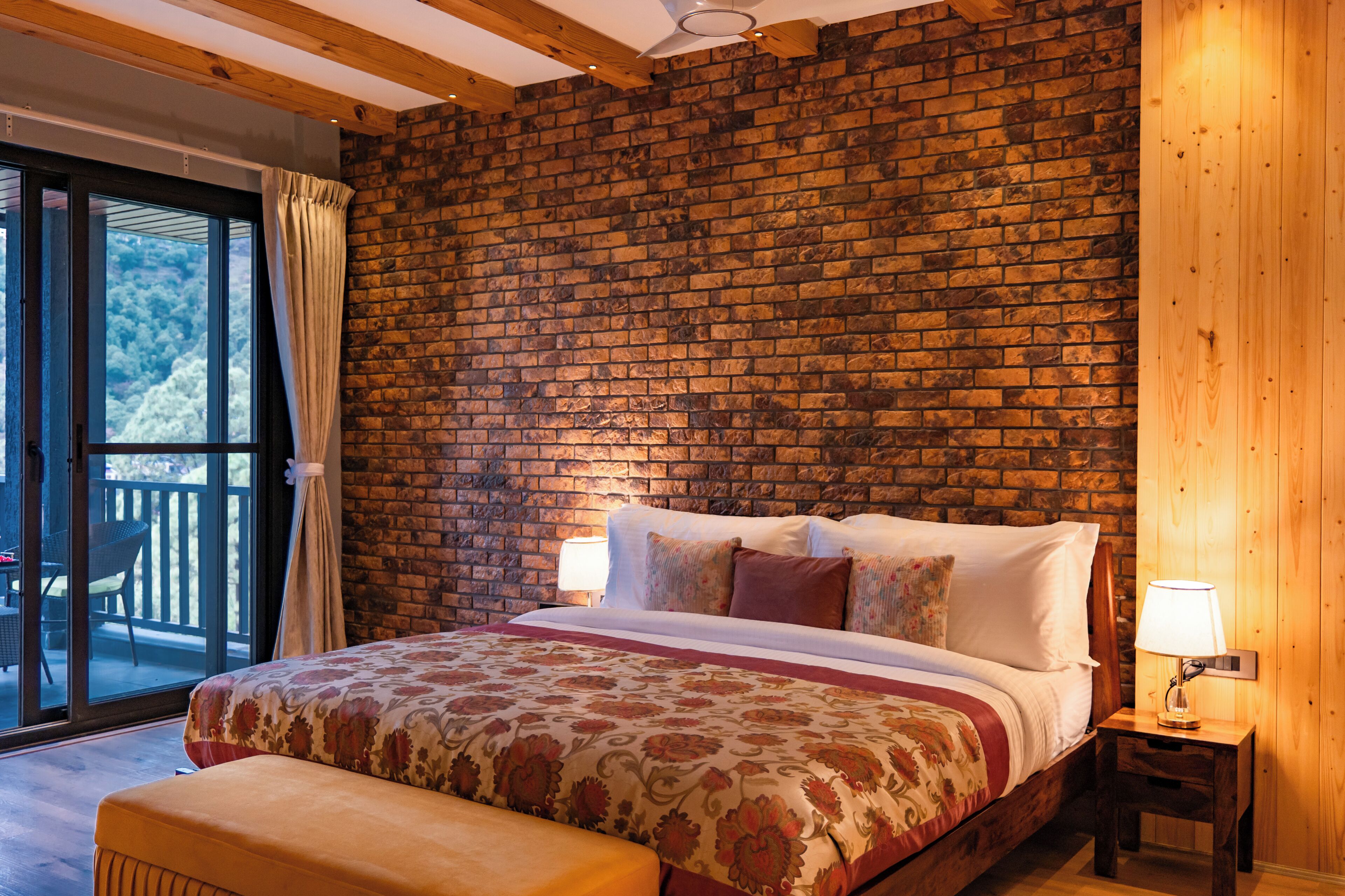 Un dormitorio con pared de ladrillo, una cama con colcha floral, una mesita de noche con lámpara y vistas a través de una puerta de vidrio.