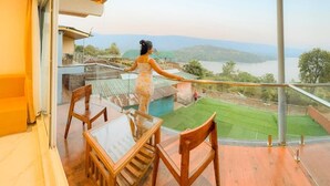 A balcony with a glass railing, a wooden table, and chairs, overlooking a green landscape and a body of water. - Nisarga Lake Resort Tapola (Mahabaleshwar)