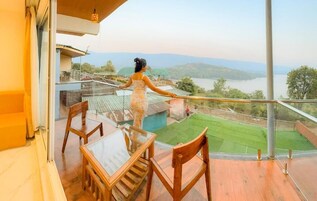 A balcony with a glass railing, a wooden table, and chairs, overlooking a green landscape and a body of water.