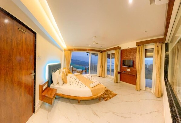 A bedroom with a curved bed, a TV, a view of the sea, and a balcony. - Nisarga Lake Resort Tapola (Mahabaleshwar)
