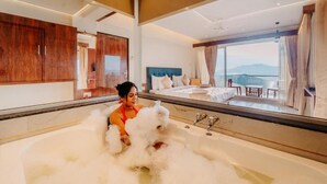 A person in a bathtub filled with bubbles, a bed with a blue headboard, and a view of the sea through large windows. - Nisarga Lake Resort Tapola (Mahabaleshwar)