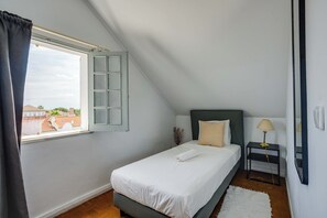 A bedroom with a bed, a window with curtains, a bedside table with a lamp, and a view of buildings outside. - Penha Single Bedroom, Hostel Idanha, Idanha-a-Nova (Idanha-a-Nova)