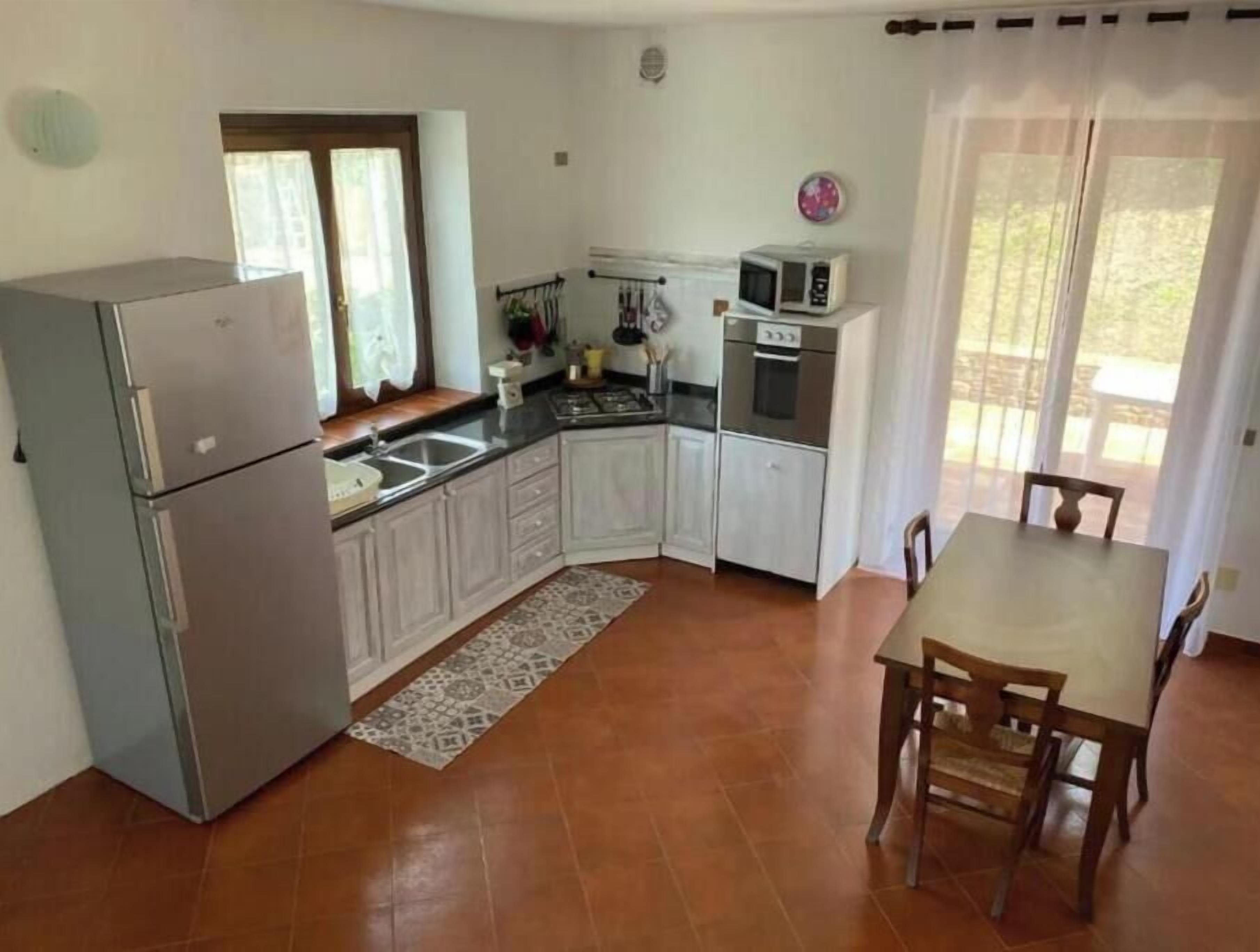 Eine kompakte Küche mit Kühlschrank, Mikrowelle, Herd und Spüle. Ein Esstisch mit Stühlen und ein Fenster mit Vorhängen.